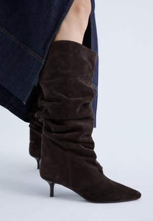 Botas flexibles de ante marrón oscuro hasta la mitad de la pantorrilla con punteras puntiagudas y tacones pequeños, combinadas con una falda oscura de mezclilla.
