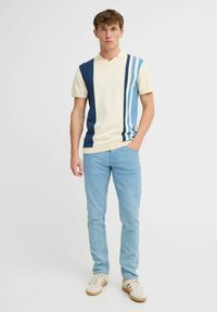 Polo a maniche corte di colore crema con strisce verticali blu, dotato di colletto. Abbinato a jeans azzurri e sneakers.