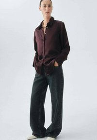 Camisa burdeos de botonadura en tejido suave, combinada con jeans de mezclilla oscuros de cintura alta. La modelo lleva un collar de perlas y zapatos en tonos neutros.