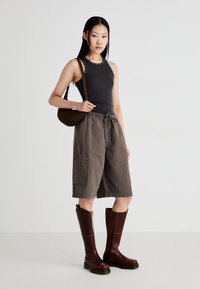 Brown knee-high boots, gray cargo shorts with side pockets, and a black sleeveless top with decorative studs. Brown shoulder bag. Plain background.