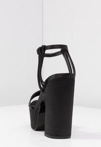 Černé sandály na vysokém podpatku s tlustou platformou, s matným textilním svrškem a štíhlými pásky kolem kotníků. Pohled zezadu zdůrazňuje blokový podpatek.