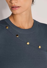 Frau trägt einen dunklen, gerippten Pullover mit einer diagonalen Reihe glänzender goldener Knöpfe über der Brust und im Kragenbereich.