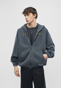 Jonge man die een donkergrijze capuchontrui met rits draagt over een zwart T-shirt en zwarte broek, kijkt opzij met één hand in zijn zak.