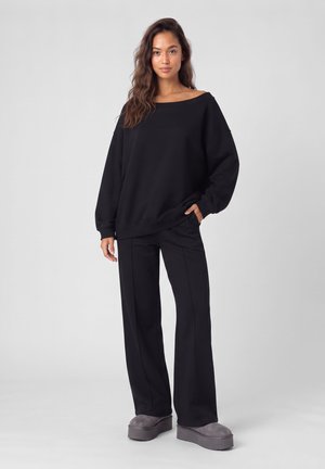 Jeune femme aux longs cheveux ondulés portant un pull noir ample, un pantalon noir à jambes larges et des chaussures compensées grises, debout devant un fond uni.