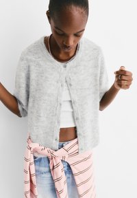 Hellgrauer, kurzärmeliger Cardigan mit einer weichen, flauschigen Textur und Knöpfen an der Vorderseite, kombiniert mit einem weißen Tanktop und einem geknoteten, rosa gestreiften Tuch.