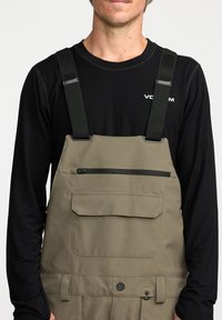 Khaki overalls met verstelbare zwarte banden, voorzien van een ritszak op de borst en details op het voorpaneel. Draag dit over een zwart T-shirt met lange mouwen.