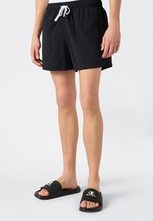 Nogi u czarnych kąpielówek z białym sznurkiem i czarnych klapkach Champion na jasnoszarym tle.