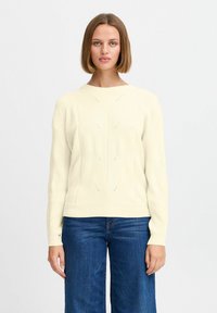 Krémový pletený svetr s kulatým výstřihem, texturovaným vzorem a ribbovanými manžetami. Slučuje se s modrými širokými džíny.