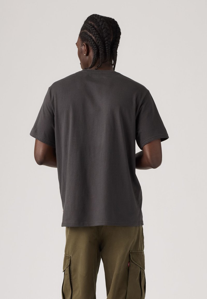 Hombre con trenzas, vestido con una camiseta lisa gris oscuro y pantalones cargo verde oliva, visto de espaldas sobre un fondo sencillo.