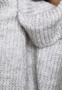 Světle šedý pletený svetr s žebrovaným rolákem, který má měkkou strukturu a pohodlný volný střih. Detaily ukazují silný pletený vzor.