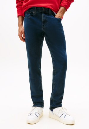 Férfi sötétkék farmerben, fehér sportcipőben és piros hosszú ujjú ingben, az egyik kezével a zsebében, fehér háttér előtt állva.