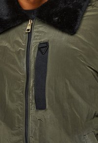 Chaqueta bomber verde oliva con un cuello de piel sintética negro, cierre de cremallera y un detalle de lazo de tela negra en el frente. Textura suave.