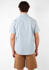 Chemise à manches courtes bleu clair avec couture au dos, ourlet arrondi et un petit patch logo près du col. Associée à un short kaki.