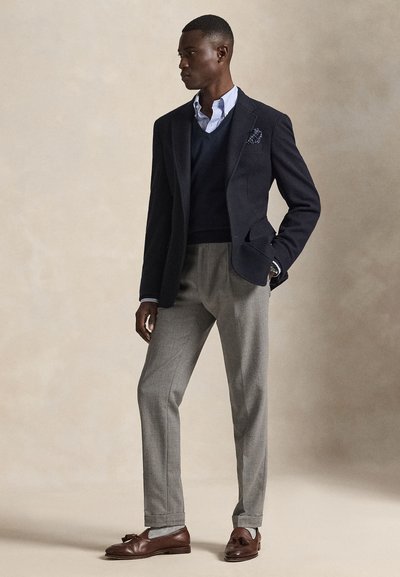 Uomo che indossa un blazer blu navy, un gilet di lana blu sopra una camicia a righe, pantaloni grigi e mocassini di pelle marrone, in piedi davanti a uno sfondo semplice.