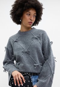 Pilkos megztos suknelės, dekoruotų kaspinų, apvaliu kaklu ir laisvu siluetu. Suderinta su juoda naginėta rankine. Matomi džinsiniai dugnai.