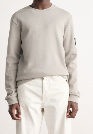 Sweat-shirt gris clair en maille gaufrée avec manches longues, col rond et poignets côtelés, associé à un pantalon de couleur crème. Détail subtil de texture.