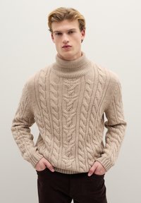Maglione dolcevita beige a trecce con motivo testurizzato e collo a coste. Il modello indossa pantaloni marrone scuro con le mani in tasca.