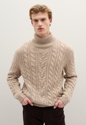 Pull à col roulé en maille torsadée beige avec un motif texturé et un col côtelé. Le mannequin porte un pantalon marron foncé, les mains dans les poches.