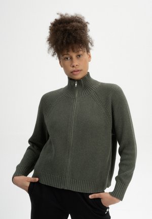 Grüner Strickpullover mit hochgeschlossenem Kragen und halbem Reißverschluss, mit geripptem Muster und Raglanärmeln. Der Saum hat einen geraden Abschluss.
