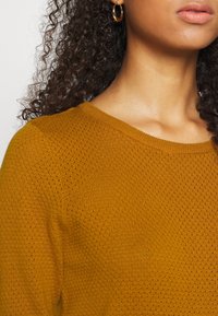 Pull en tricot jaune moutarde avec un col rond, présentant un motif texturé, accompagné de boucles d'oreilles créoles dorées.