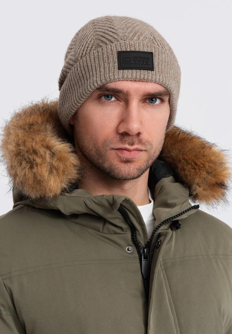 Gorro de punto color beige con textura acanalada y una etiqueta de marca negra. Llevado con una parka verde con capucha forrada de piel.