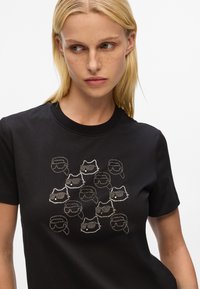 Schwarzes Baumwoll-T-Shirt mit einem Grafikdesign, das mehrere Katzenfaces in Weiß mit Sonnenbrillen zeigt, angeordnet in einem verspielten Clusterdesign.