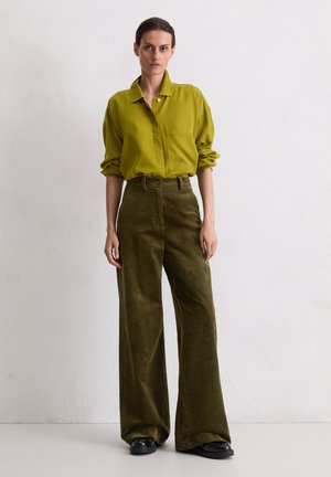 Olijfgroene overhemdbloes met een zak, gecombineerd met brede corduroy broeken in een donkerder groen, gedragen met zwarte schoenen. Glad textuur van de stof.