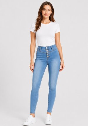 Kvinde iført en hvid t-shirt med korte ærmer, lyseblå jeans med høj talje og flere knapper samt hvide sneakers, stående på grå baggrund.