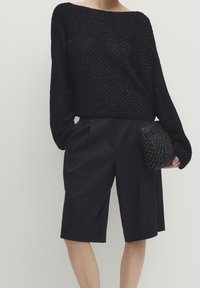 Maglione nero lavorato a maglia con vestibilità ampia e scollatura larga, abbinato a pantaloni corti neri su misura. Accompagnato da una clutch nera texture.