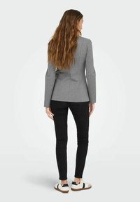 Femme aux longs cheveux ondulés portant un blazer gris ajusté, un pantalon noir slim et des baskets blanches, debout, de dos, sur un fond uni.