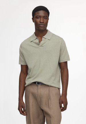 Junger Mann mit neutralem Gesichtsausdruck, trägt ein hellgrünes Polohemd mit kurzen Ärmeln und beige Hosen mit schwarzem Gürtel.