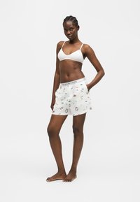 Γυναίκα στέκεται ξυπόλυτη, φοράει λευκό μπραλετί και λευκά μποξεράκια με πολύχρωμα σχέδια και λογότυπο στη μέση, τα χέρια στις τσέπες.