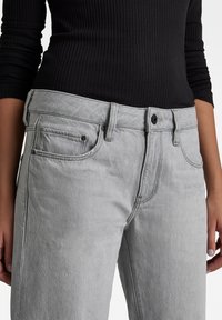 Pessoa vestindo jeans de ganga cinzentos claros com bolsos frontais e uma blusa de mangas longas preta de canelado, com as mãos ao lado.