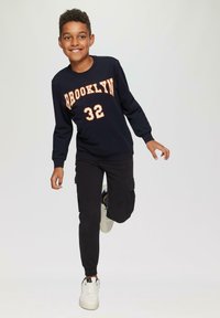 Zwarte crewneck sweatshirt met "BROOKLYN 32" in oranje gedrukt. Gecombineerd met zwarte cargobroek en witte trainers. Jongeman in een speelse pose.