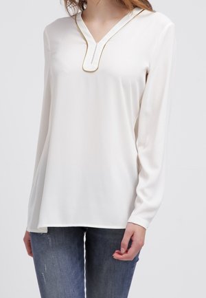 Blouse blanche à manches longues avec un col en V et un accent doré le long de l'encolure, fabriquée à partir d'un tissu lisse, avec une coupe ample.