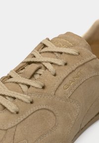 Gros plan sur une basket en daim beige avec des lacets épais couleur crème et le logo doré "GANT" sur le côté et la languette.