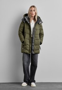 Olivgrön puffjacka med en mönstrad svart och vit huva, dragkedja fram, två sidofickor och justerbara snörningar i nederkant.