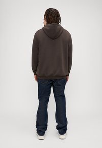 Brun hettegenser med snor, med en myk tekstur. Kombinert med mørkeblå jeans og hvite joggesko, sett fra ryggen.