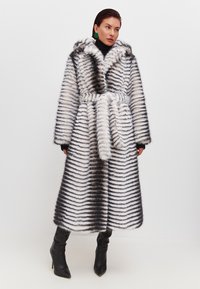 Cappotto in pelliccia sintetica con cappuccio, caratterizzato da strisce bianche e nere. Maniche ampie, vita con cintura e design di lunghezza media. Indossato sopra un dolcevita.