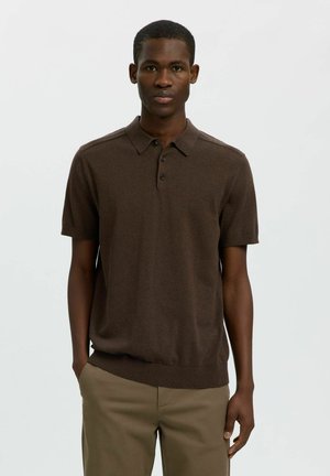 Jeune homme portant un polo marron à manches courtes et un pantalon beige, debout avec une main dans la poche sur un fond uni.
