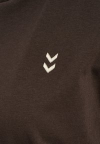 Brun bomuldsstof med et lille, hvidt, broderet logo bestående af to chevron-former, placeret på venstre side.