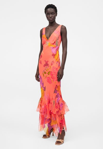 Γυναίκα που φοράει ένα κοραλί αμάνικο μακρύ φόρεμα με ροζ, πορτοκαλί και μοβ λουλουδένια σχέδια και βολάν στο τελείωμα, συνδυασμένο με μπεζ πέδιλα με τακούνια.
