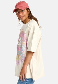Light beige oversized t-shirt with a colorful graphic design featuring clouds and flowers. Paired with a pink baseball cap.