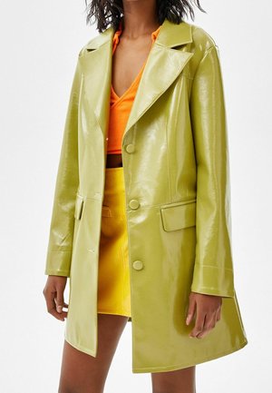 Femme portant un manteau boutonné vert clair brillant sur un haut orange et une jupe jaune, debout devant un fond uni.