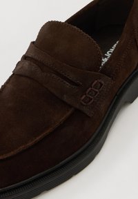 Kestenjasta odsuđena loafer cipela s prošivenim detaljima, zaobljenim vrhom i teksturiranom crnom gumenom petom. Ima dekorativnu traku preko prednjeg dijela.