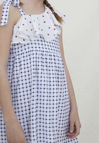 Abito a quadri bianco e blu navy con scollo arricciato bianco decorato da motivi a cuore rossi, fissato con fiocchi sulle spalle.