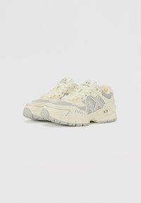 Baskets de sport beige clair avec des accents gris, présentant un dessus en maillage, un col rembourré et une semelle en caoutchouc texturé. Logo sur la languette et les côtés.