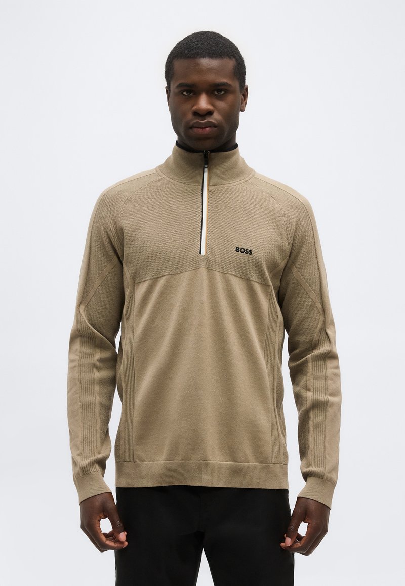 Άνδρας φοράει μπεζ πουλόβερ BOSS με φερμουάρ στο στήθος, με υφασμένες πλάκες και ριμπ μανίκια, στέκεται μπροστά σε ένα απλό φωτεινό φόντο.