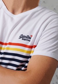 Vit T-shirt med horisontella ränder i rött, gult och marinblått, med en liten broderad "SuperDry"-logotyp i röd färg på bröstet. Mjuk bomullstextur.