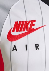 Nike Air -paita, jossa valkoinen runko harmailla ohuilla raitakuoseilla, punainen kirjailtu logo ja mustat korostus-yksityiskohdat olkapäillä.